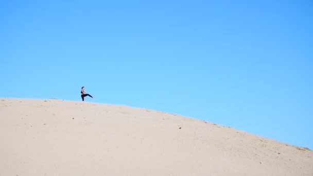 身穿黑色运动服的女孩 练习抬腿 完成踢腿动作 用影子打斗 训练打斗 在炎热的阳光下 在荒芜的海滩上 在蓝天的映衬下 — 图库视频影像