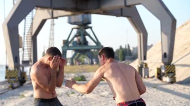 Çıplak, çıplak torsolar, kutusu, uygulama ile genç atletik erkek teknik grev, yakalama, mücadele, büyük kargo Vinçler, yakındaki bir kargo limanda kumlu bir plaj üzerinde gündoğumu, yaz aylarında.