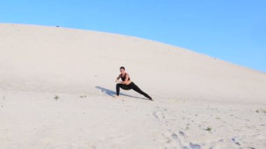 Siyah bluzlu ve taytlı atletik bir kız sicim pozisyonunda, ıssız bir sahilde jimnastik sicimde oturuyor, sıcak güneşin altında, mavi bir gökyüzüne karşı.