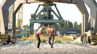 Çıplak, çıplak torsolar, kutusu, uygulama ile genç atletik erkek teknik grev, yakalama, mücadele, büyük kargo Vinçler, yakındaki bir kargo limanda kumlu bir plaj üzerinde gündoğumu, yaz aylarında.