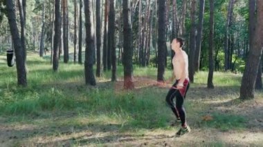 Çıplak, çıplak gövdeli, atlama halatıyla genç bir atletik, kauçuk bir iple güç egzersizleri yapıyor, yaya geçidi elementi. Ağır çekim. Çam ormanlarında, yazın, güneş ışınlarında