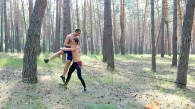 Çıplak vücutlu genç atletik erkekler, vuruş tekniğini uygularlar, yakalarlar, dövüşürler, lastik bir iple, ağır çekimde. Çam ormanlarında, yazın, güneş ışınlarında.