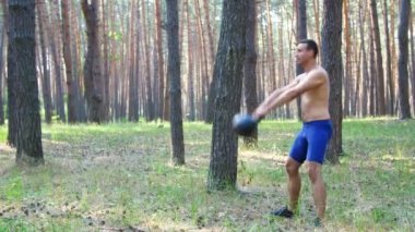 Çıplak, çıplak bir gövdesi olan genç atletik bir adam, çapraz formda olan ağırlık egzersizleri yapıyor. Ağır çekim. Çam ormanlarında, yazın, güneş ışınlarında.