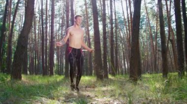 Çıplak, çıplak gövdeli, atlama halatıyla genç bir atletik, kauçuk bir iple güç egzersizleri yapıyor, yaya geçidi elementi. Ağır çekim. Çam ormanlarında, yazın, güneş ışınlarında