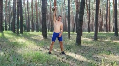 Çıplak, çıplak bir gövdesi olan genç atletik bir adam, çapraz formda olan ağırlık egzersizleri yapıyor. Ağır çekim. Çam ormanlarında, yazın, güneş ışınlarında.
