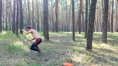 Genç atletik biri bir çıplak, çıplak gövde ile güç egzersizleri bir kauçuk ip, crossfit öğesi ile gerçekleştirir. Ağır çekim. Çam ormanı içinde yaz, güneş ışınlarının.