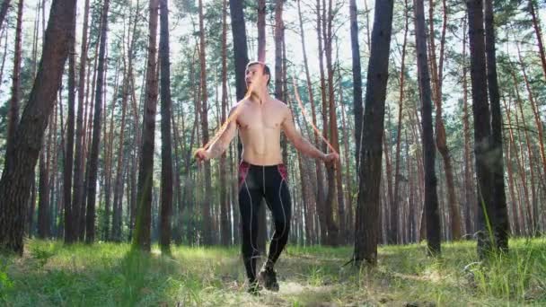 一个年轻的运动员 身材矮小 赤身裸体 跳着绳子 用一根橡皮绳 一个交叉配合的元素进行力量训练 慢动作在松林里 在阳光下 — 图库视频影像