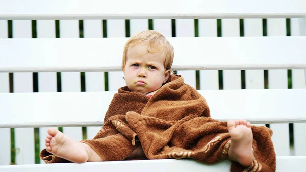 a little one-year-old girl, wrapped in a towel, with a dirty face, lonely sitting on a swing in the garden, in the summer. She has a sad look. She wants to sleep. High quality photo