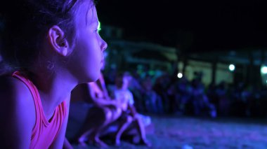Yaz, gece, Soffits 'in ışınlarında, yedi yaşında bir kız, açık hava tiyatrosunda gece konseri izliyor, tatilde. Arka planda bir sürü çocuk var. Yüksek kalite fotoğraf