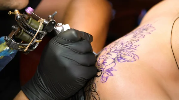 close-up, in a tattoo salon , a specialist is doing a tattoo on womans back, black paint floral ornament. a man works in special black gloves, on special equipment. High quality photo