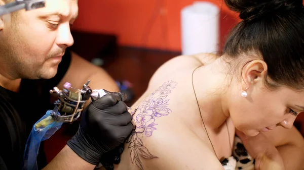 in a tattoo salon , a specialist is doing a tattoo on womans back, a floral ornament. a man works in special gloves, on special equipment. drawing is done with black paint. High quality photo