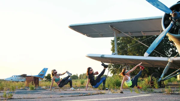 Beautiful, athletic, young women in sunglasses, in tights, perform synchronously different strength exercises, jumps, push-ups, lifting legs.on an abandoned airfield, near plane. High quality photo