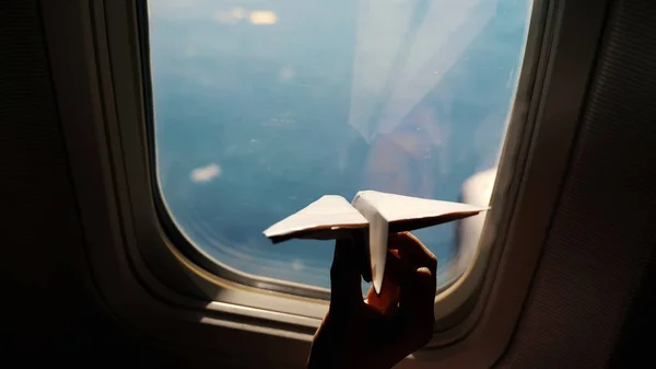 close-up. Silhouette of a childs hand with small paper plane against the background of airplane window. Child sitting by aircraft window and playing with little paper plane. during flight on airplane