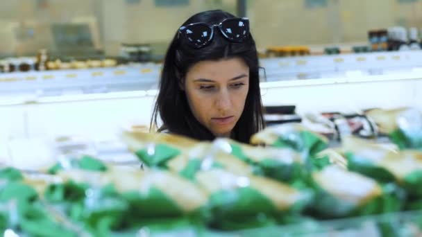 女性はスーパーで食料品を選ぶ ポートレート ブルネットの女性は 食料品部門のスーパーマーケットの棚に横たわる製品を慎重に調べています 食品を購入する — ストック動画