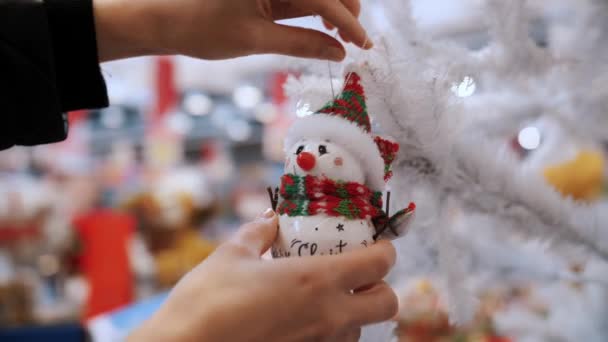 クリスマスアクセサリー クローズアップ 手は店の白い人工的なクリスマス ツリーの面白い雪だるまの形でクリスマスのおもちゃ 装飾を掛けます 公正な スーパーマーケットまたはクリスマスの装飾 — ストック動画