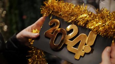 Yeni yıl 2024 plakalı altın rakamlar. Noel aksesuarları. Yakın plan. Kadın, dükkanda üzerinde 2024 yazısıyla bir Yeni Yıl tabelası tutuyor. Mutlu yıllar 2024 alışverişler. Noel Fuarı