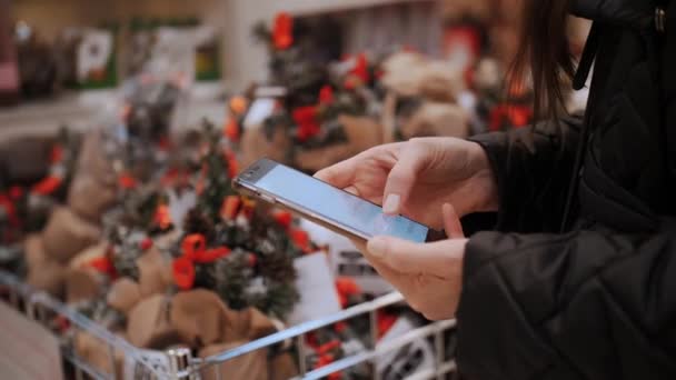 携帯電話を使って クリスマスの装飾 ショッピング クローズアップ スマートフォンでタイピングする顧客は ギフトを選択しながら フェア スーパーマーケット ショッピングモールでのお祝いのアクセサリー — ストック動画