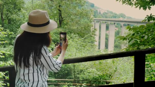 彼女の携帯電話でアルプスを捕まえた帽子をかぶった女性 バックグラウンドでは 車の橋 公園の夏に 高品質の4K映像 — ストック動画