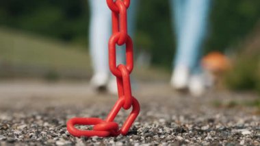 Sallanan kırmızı metal zincir. Yakın plan. Zincir halkaları. Yoldaki asfaltta asılı kırmızı bir zincir var. Arka planda beyaz spor ayakkabılarla yürüyen kadın bacakları var.