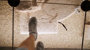 İlk bakış açısı. Cam altında arkeolojik kazılar müzesi. Tarihi eserler. Sağlam bir cam yüzeyde yürüyen bir turistin ayaklarının en üst görüntüsü. Yüksek kalite 4k görüntü