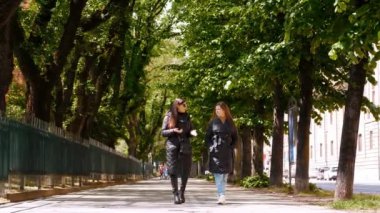 İki kadın, uzun bir ağacın gölgesinde, pitoresk bir caddede yürüyor. Bir anlık dostluk ve rahatlama.