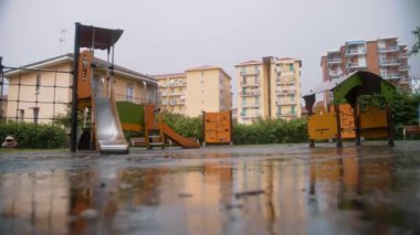 Yağmur sonrası banliyö ortamında ıslak bir oyun parkının canlı bir tasviri, su birikintileri, renkli oyun ekipmanları ve çevresindeki konut binaları yansıtıyor