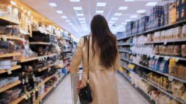 Arkadan resmedilen genç bir kadın, süpermarket koridorlarında ürünlere göz atıyor, günlük tüketici faaliyetlerini ve perakende çevre nüanslarını vurguluyor
