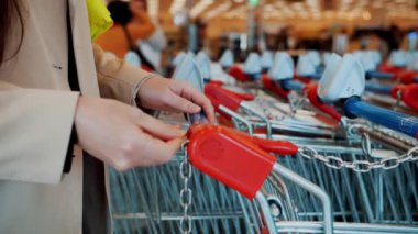 Yoğun bir süpermarketten alışveriş arabası alan bir kadın. Bozuk para yerleştirir ve dükkandan alışveriş arabasını alır. Bakkalda Bir Alışveriş Arabası Kiralamak İçin Bir Bozuk Para Kullanan Kadınlar