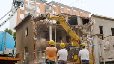 Güvenlik kaskı ve yansıtıcı yelek giyen üç inşaat işçisi, çok katlı bir binanın yıkımına tanık oluyor. Hidrolik öğütücüyle donatılmış büyük sarı bir kazıcı aktif olarak çalışır.