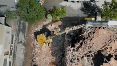 Sarı bir kazıcının yukarıdan aşağıya doğru görüntüsü eski bir tuğla binayı yıkıyor, inşaat faaliyetleri ile çevre çevre çevre arasındaki zıtlığı yakalıyor.