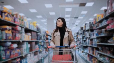 Trençkot giymiş bir kadın, bir alışveriş arabasını süpermarket koridoruna itiyor, çeşitli ürünleri dikkatlice düşünüyor. Odaklanmış ifadesi tüketici davranışlarını yansıtıyor.