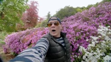 Şaşırmış bir adam, serin, canlı rhododendron çalılarının büyüleyici arka planına sahip bir selfie çeker. Parkta pembe ve mor çiçeklerin karışımını sergiler.