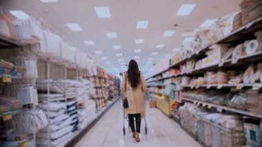 Perakende mağazasında ev eşyalarıyla dolu bir koridorda yürürken bir kadın arkadan görülüyor. Sıradan giyinmiş ve bir çanta taşıyor. Temiz ve düzenli bir ortamda alışveriş yapıyor.