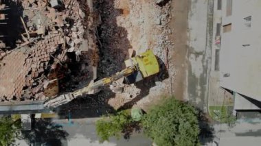 Şehir yıkım alanındaki molozların arasında, bozulmamış binalarla çevrili sarı bir kazıcının detaylı görüntüsü.