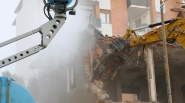 Güçlü bir hidrolik kazıcı binanın bir kısmını enkaz ve tozla yıkıyor ve tipik bir kentsel yenilenme sahnesi sergiliyor.