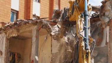 Zırhlı bir kazı aracı, bir apartman dairesini sökerken yıkıntıların arasında duruyor. Arkaplanda modern bir bina tarafından vurgulanan inşaat sahasının etrafına demir parçaları ve enkaz parçaları saçılmış.