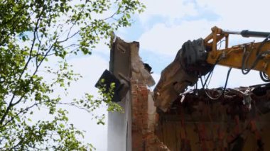Zarar görmüş tuğla bir binanın kalıntılarını parçalayan sarı kazıcının detaylı bir görüntüsü, açık mavi gökyüzüne karşı yeşil yapraklar arasında.