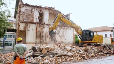 Güçlü bir kazıcı, eski bir binayı enkaz gibi yıkıyor. Güvenlik ekipmanları kullanan bir işçi tarafından yakından denetleniyor ve kentsel gelişimi gösteriyor.