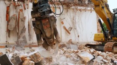 Toz ve enkaz bulutlarıyla çevrili tuğla bir binayı yıkan yıkım pençeli bir hidrolik kazı makinesinin detaylı bir görüntüsü.