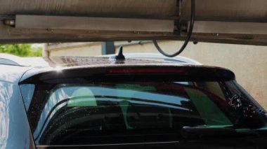 Arabanın ön cam ve çatıda su damlacıklarıyla yıkandığı detaylı bir fotoğraf, temizlik sırasında güneş ışığı. Otomatik araba kurutma.