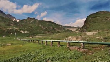 Berrak bir gökyüzünün altındaki yemyeşil vadide, engebeli dağ manzarasıyla çevrili taştan sütunlar üzerinde önemli bir boru hattı bulunuyor. Doğa ve endüstriyel temalar için ideal