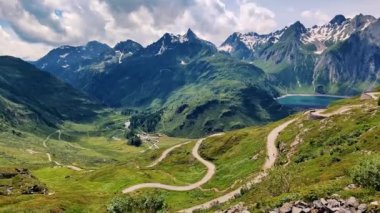 Karla kaplı dağların altında kıvrımlı yeşil çayırlarda kıvrılan kıvrımlı yolların yer aldığı bir dağ manzarasının nefes kesici güzelliğini yakalayın.