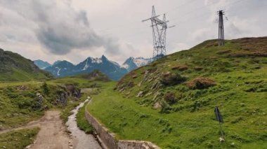 Çimenli verimli tarlalar ve karla kaplı görkemli dağlara doğru uzanan toprak bir yol dramatik bulutlu gökyüzü altındaki elektrik hatlarıyla çevrelenmiş doğa ve altyapı temaları için mükemmel.