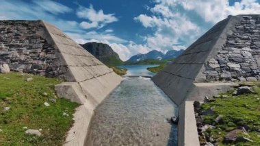 Görüntüde, yemyeşil ve engebeli dağlarla çevrili sakin bir alp gölüne uzanan berrak, dar bir dereyi çerçeveleyen iki heybetli beton yapı yer alıyor.