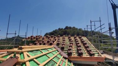 Fotoğraf, açık mavi bir gökyüzünün altında seramik işçiliğine odaklanan bir konut çatısının devam eden yapısını gösteriyor. Ahşap iskele karmaşık yapıyı destekler