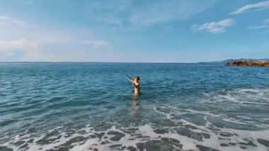 Neşeli bir kadın, beline kadar berrak mavi okyanus sularına batmış, parlak gökyüzünün altında, zevkle yükselmiş, özgürlüğü ve yaz neşesini sembolize eden,