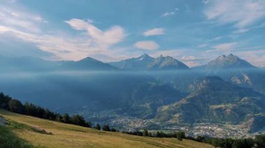 Açık mavi gökyüzünün altındaki görkemli dağlarla çevrili yeşil bir vadinin nefes kesici manzarası, doğa ve seyahat temaları için mükemmel.