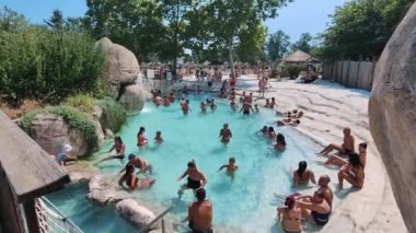 Çeşitli yaşlardan turistlerin güneşli bir günün tadını çıkardığı canlı tropikal yüzme havuzu sahnesi. Yaz tatilinin bu ideal tasviri insanların yüzdüğü, güneşlendiği ve sosyalleştiği gösteriler.