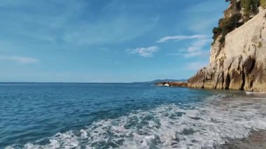 Geniş mavi gökyüzü altında berrak turkuaz sular, yumuşak dalgalar ve engebeli uçurumlar içeren kıyı manzarası, arka planlar veya seyahat temalı projeler için idealdir.