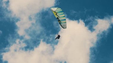 Yalnız bir paraglider gökyüzünde yükselir, ustaca manevra yaparak renkli bir gölgelik oluşturur kabarık beyaz bulutlara ve koyu mavi bulutlara karşı.